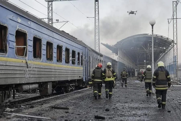 Пожарные идут мимо повреждённого пассажирского поезда по засыпанной обломками платформе; над вокзалом виден дым.