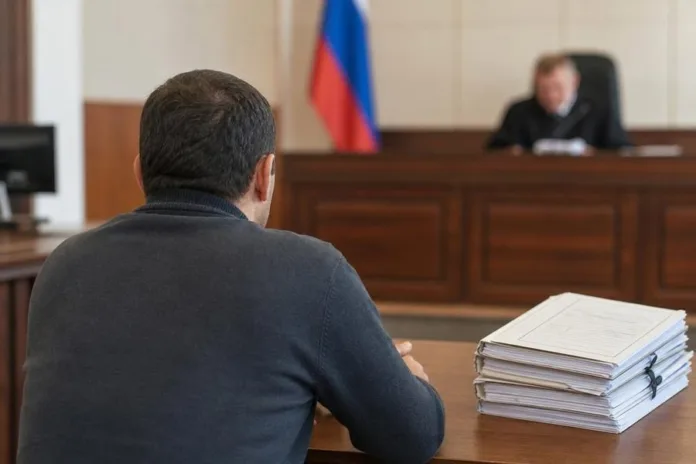 Мужчина сидит за столом напротив судьи; рядом лежит стопка документов, в зале виден российский флаг.