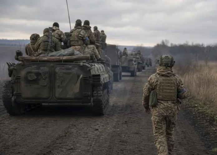 Военнослужащий идёт по просёлочной дороге рядом с колонной бронетранспортёров, на которых сидят другие военные.