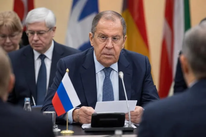 Мужчина в очках сидит за столом с документом и микрофоном; перед ним российский флаг, на заднем плане — участники встречи и флаги.
