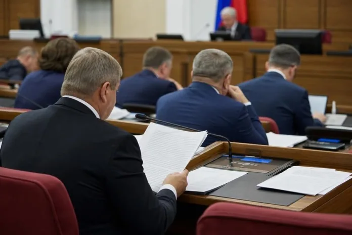 Мужчины в деловых костюмах сидят за столами в зале заседаний, один читает документ.