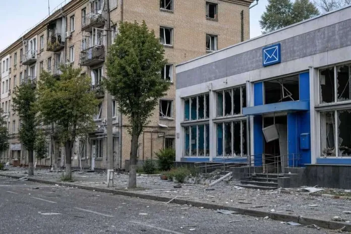 Повреждённое здание почтового отделения с выбитыми окнами стоит у многоэтажного дома; на тротуаре разбросаны обломки.