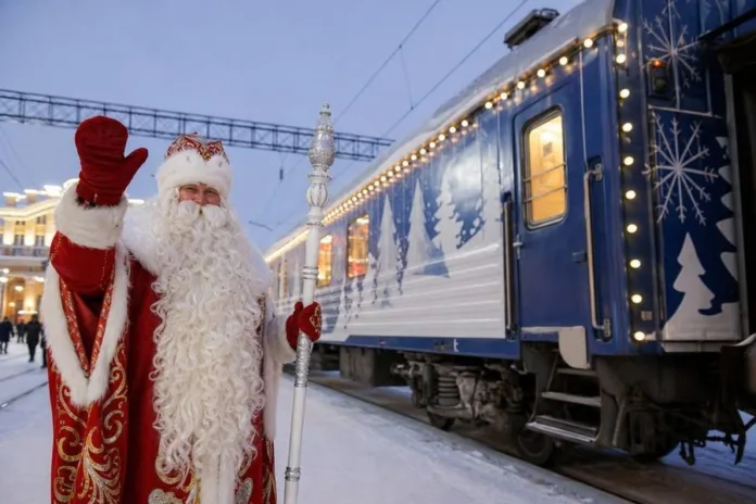 Мужчина в костюме Деда Мороза машет рукой рядом с украшенным огнями поездом на зимней платформе.