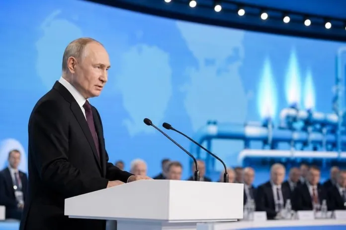 Владимир Путин выступает за белой трибуной с микрофонами перед сидящими в зале людьми.