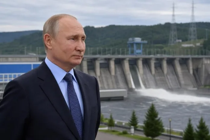 Владимир Путин стоит на фоне плотины с открытым водосбросом; вокруг видны лесистые холмы и линии электропередачи.