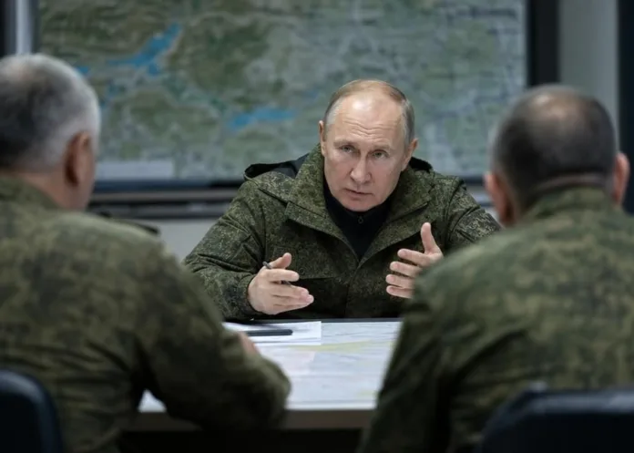 Владимир Путин в военной форме разговаривает за столом с двумя мужчинами; на стене видна карта.