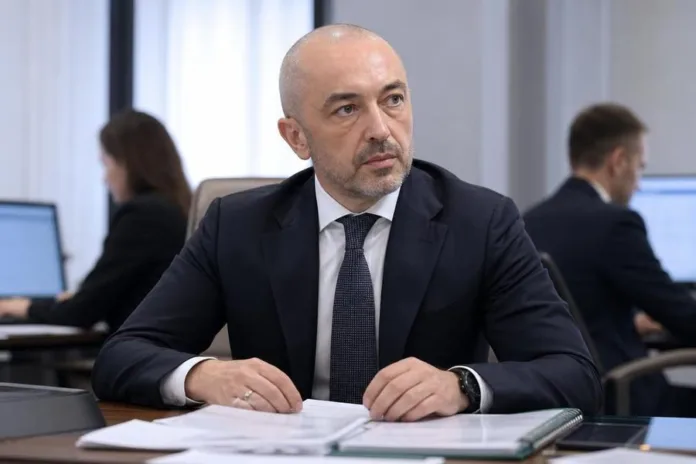 Мужчина в деловом костюме сидит за столом с документами в офисе; на заднем плане работают сотрудники.