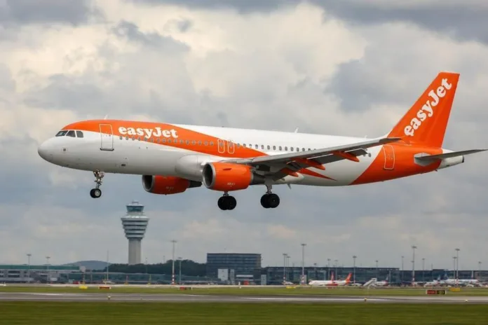 Пассажирский самолёт easyJet с выпущенными шасси летит над взлётно-посадочной полосой аэропорта; на заднем плане видна диспетчерская вышка.