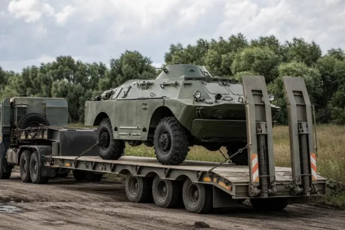 Бронетранспортёр закреплён на низкорамном прицепе грузовика на обочине среди деревьев.