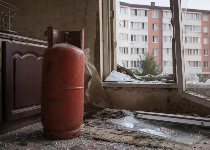 В комнате стоит красный газовый баллон; у разбитого окна на полу лежат осколки стекла.