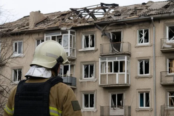 Пожарный в защитной экипировке смотрит на многоэтажный жилой дом с повреждённой крышей и разбитыми окнами.