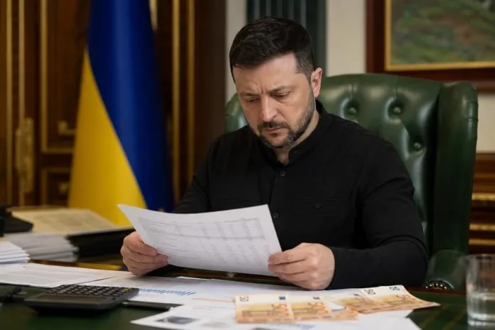 Владимир Зеленский сидит за столом и читает документ; перед ним лежат бумаги и банкноты, позади виден украинский флаг.