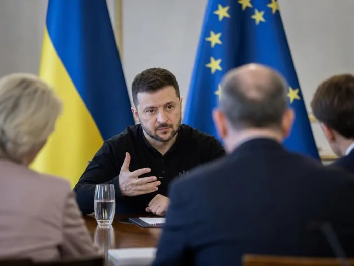 Владимир Зеленский разговаривает с людьми за столом на фоне флагов Украины и Евросоюза.